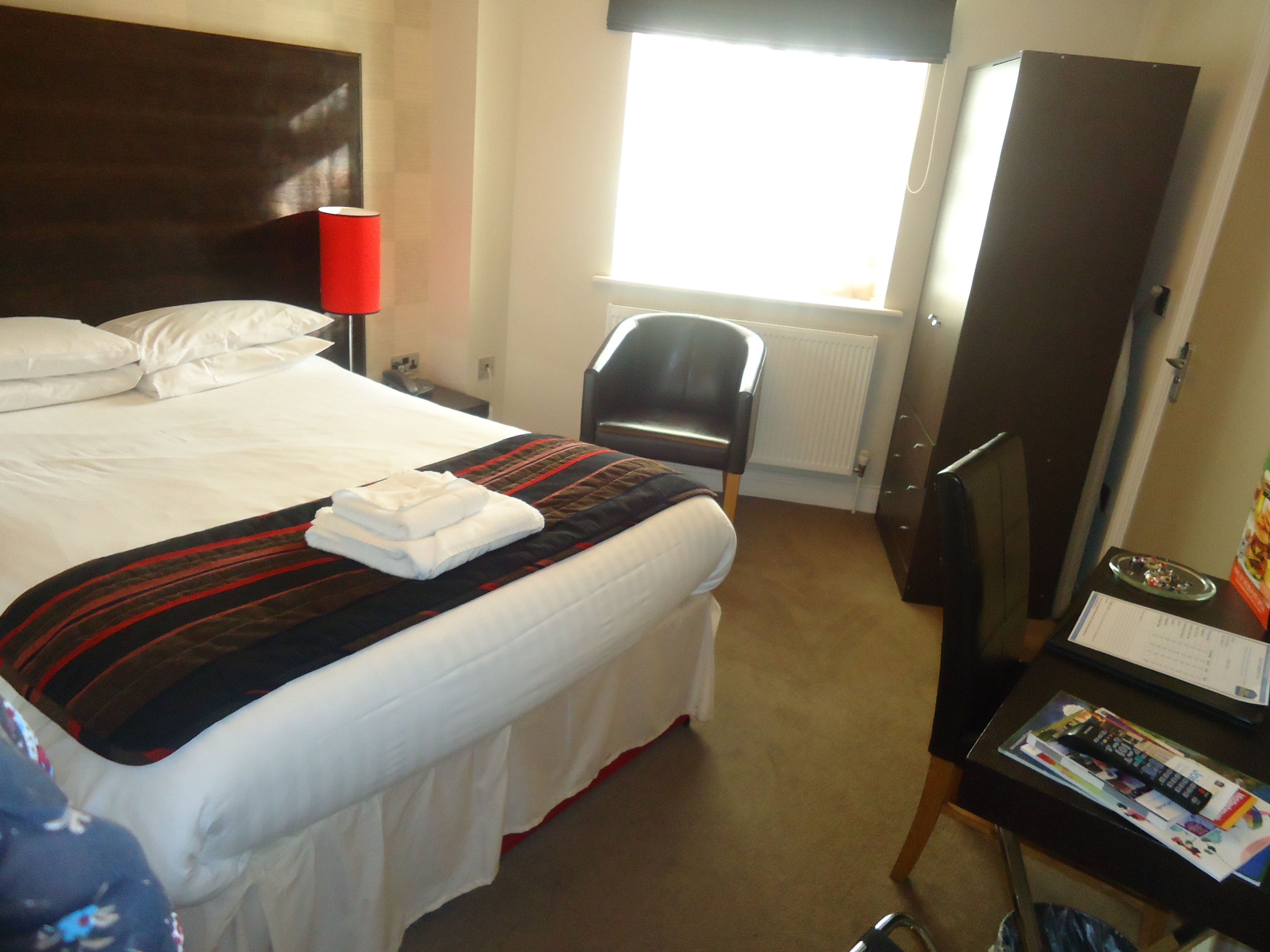 Ein Hotelzimmer mit einem ordentlich bezogenen Bett, einem Sessel, einem Schreibtisch mit Computer und einem Fenster mit Jalousien.