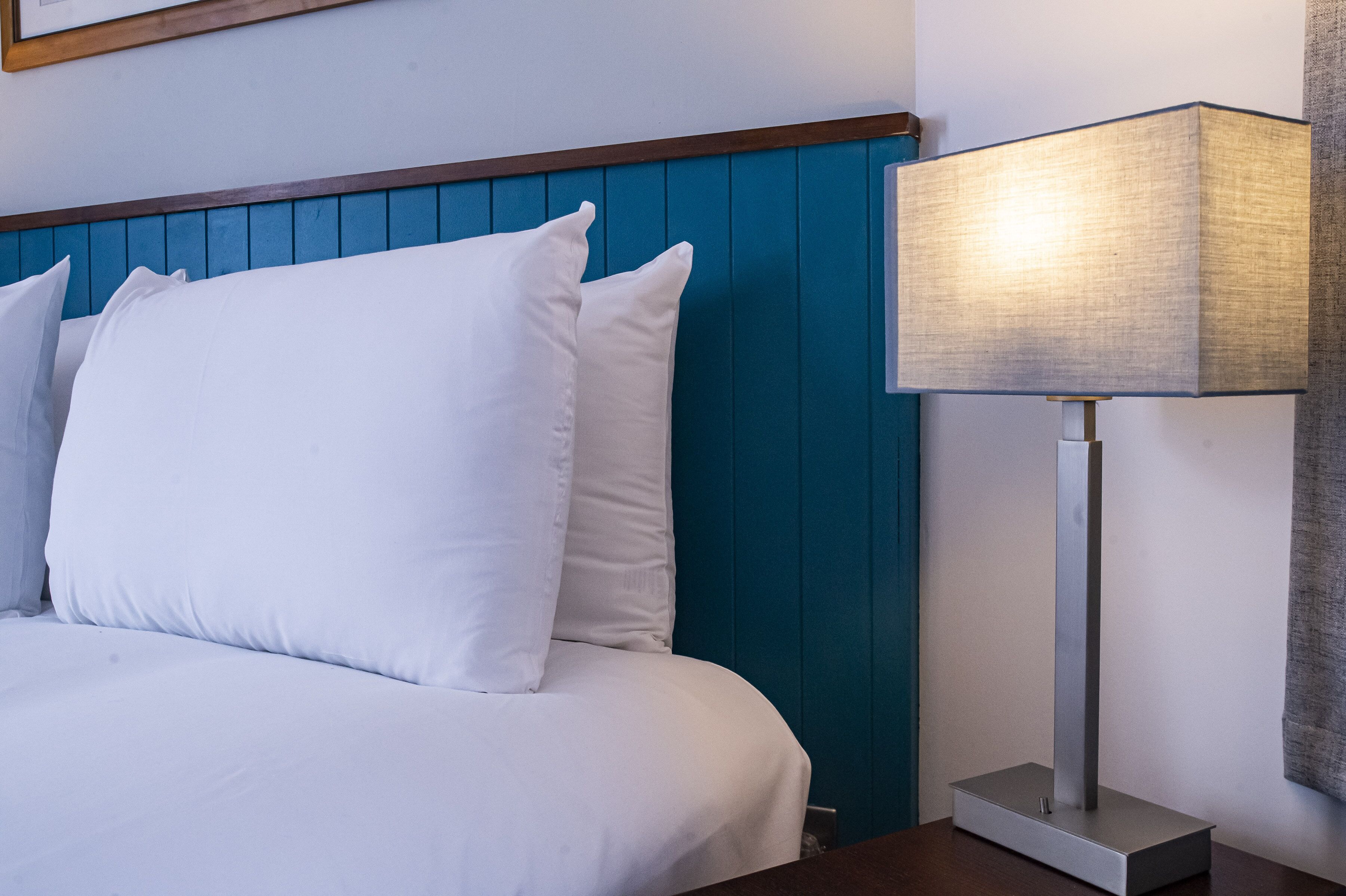 Un lit bien fait, avec du linge de lit blanc et des oreillers, une tête de lit bleue et une lampe de chevet moderne posée sur une table de nuit en bois.