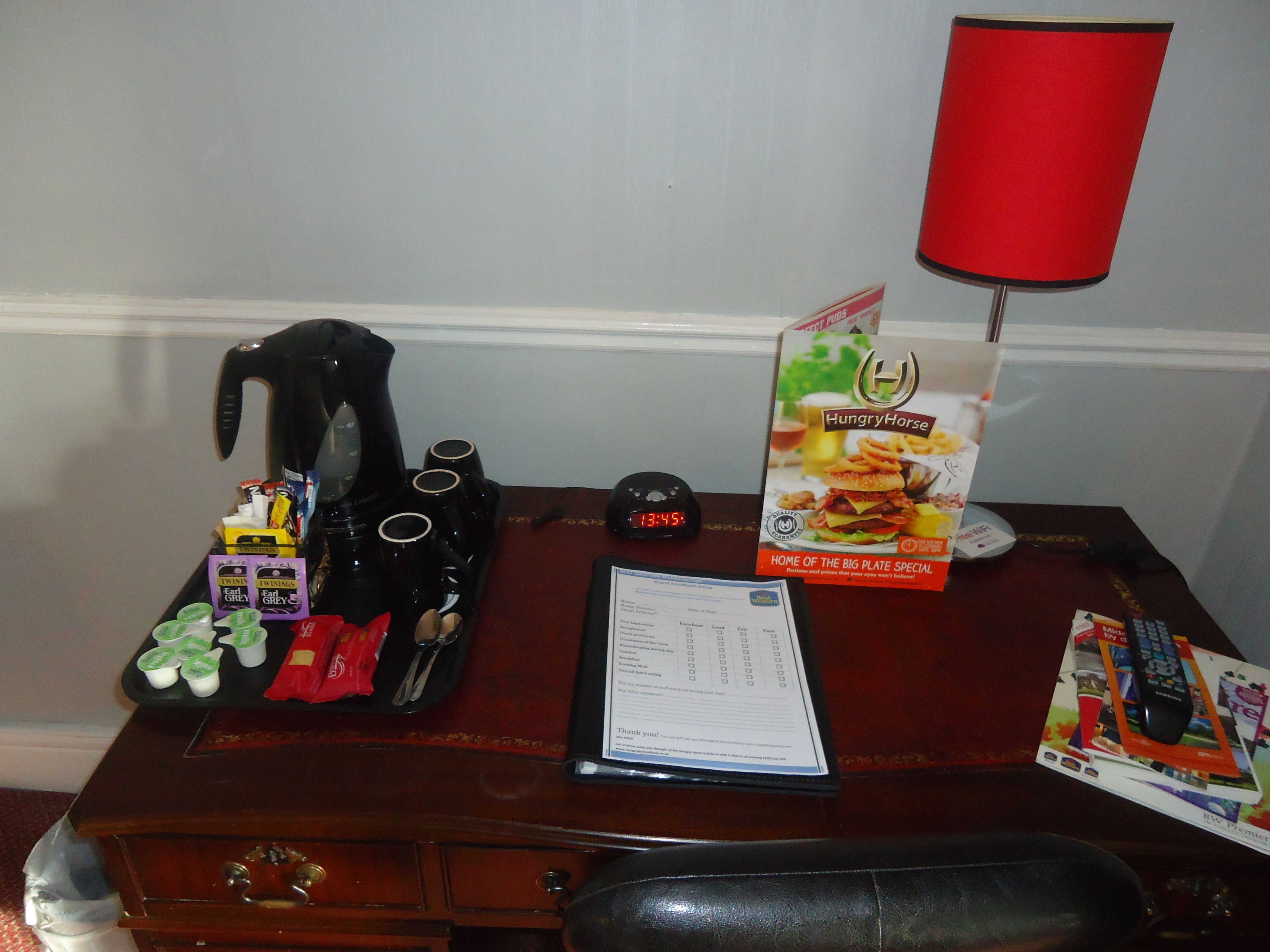 A hotel desk with a coffee maker, cups, a menu, and a red lamp.