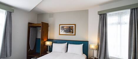 A hotel room with a bed, two bedside tables, a nightstand, a framed picture on the wall, and a window with curtains.