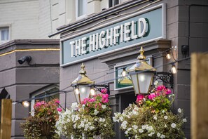 A building with a sign reading "THE HIGHFIELD" and a "DINING" sign below it. There are hanging flower baskets and two ornate street lamps.