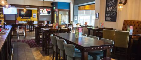 A restaurant with multiple tables and chairs, a bar area, and a television screen displaying a sports event.