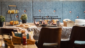 Un comedor con una mesa de buffet, que incluye una cesta de frutas de tres niveles, una selección de pasteles y una variedad de panes.