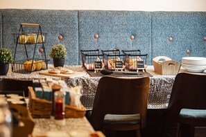 A dining area with a buffet setup, including a three-tiered fruit basket, a selection of pastries, and a variety of bread.