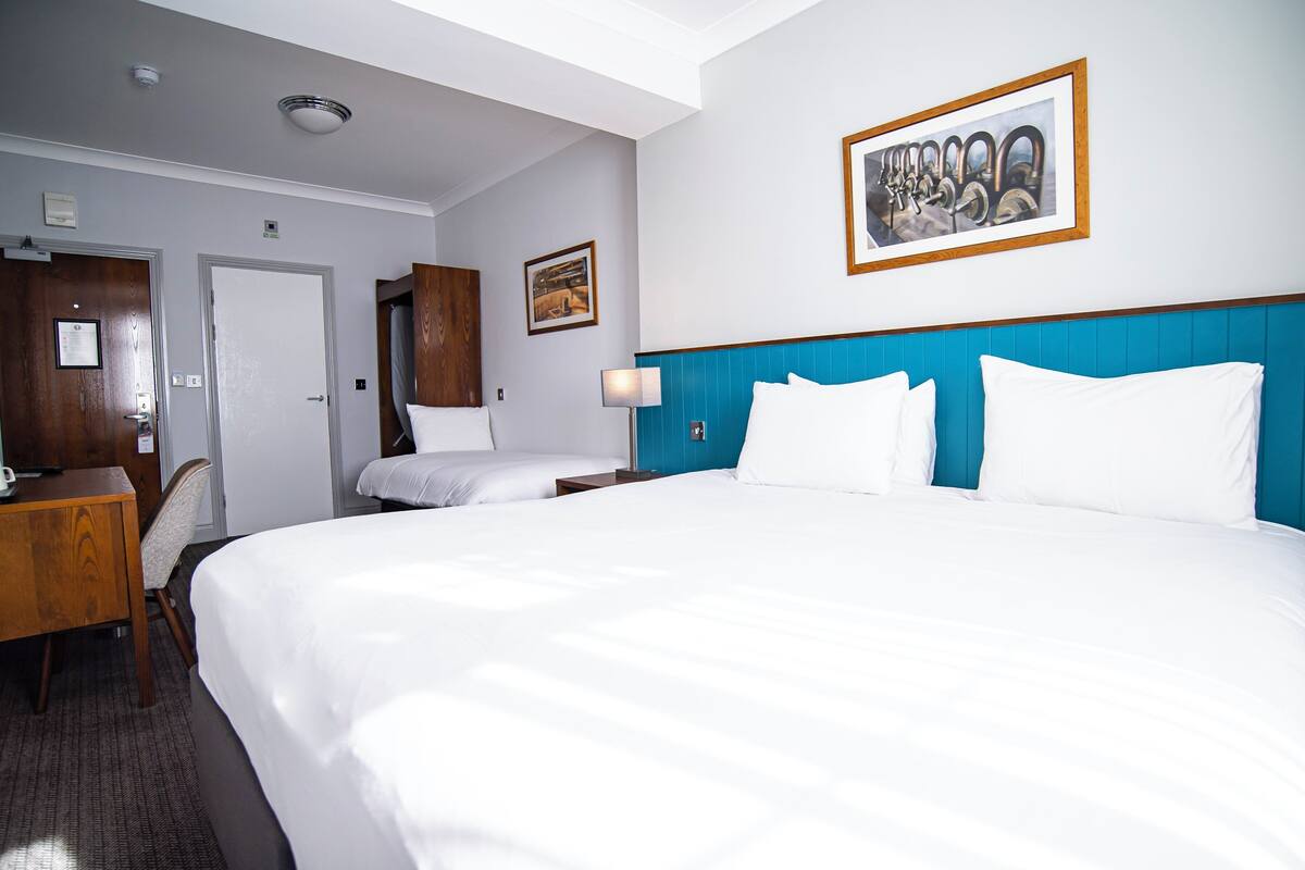 A hotel room with a large bed, a desk, and a chair. There is a door, a framed picture on the wall, and a light fixture above the bed.
