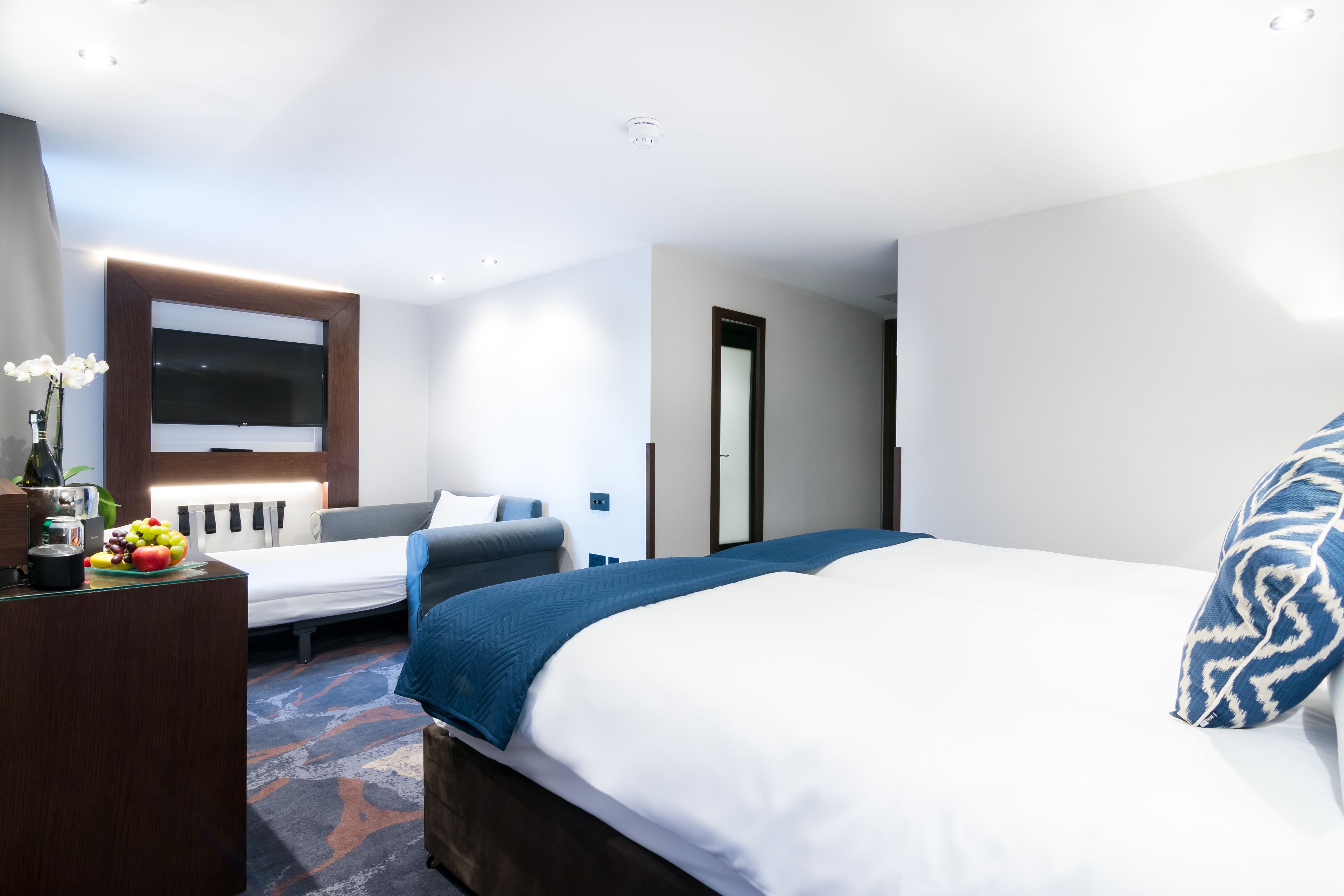 Ein modernes Hotelzimmer mit einem großen Bett, einem Schreibtisch mit Obstschale, einem Fernseher und einem Spiegel.
