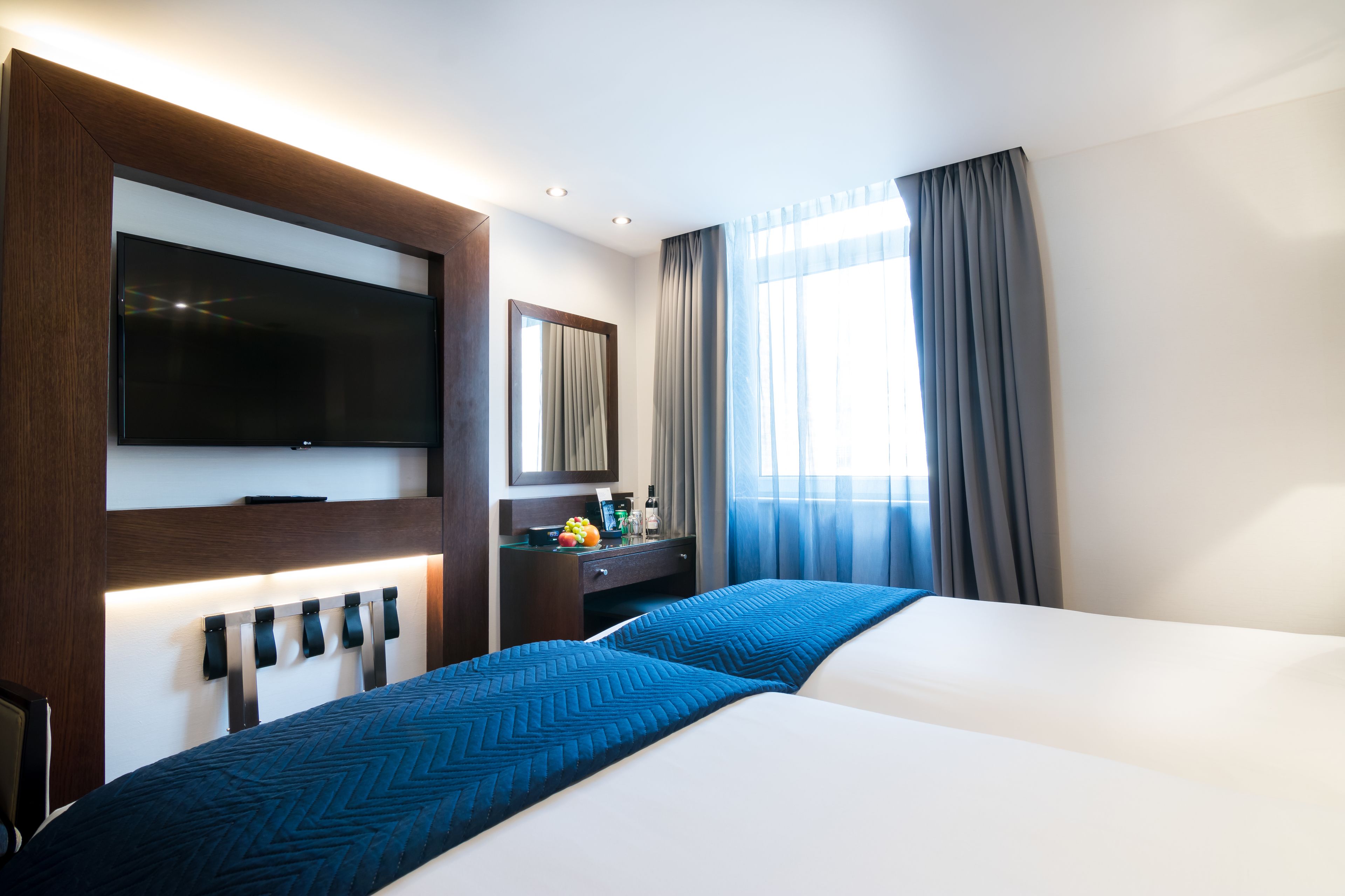 A hotel room with two beds, a flat-screen TV mounted on the wall, a desk with a mirror, and a window with curtains.