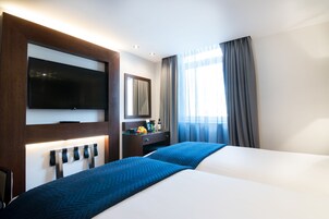 Ein Hotelzimmer mit zwei Betten, einem an der Wand montierten Flachbildfernseher, einem Schreibtisch mit Spiegel und einem Fenster mit Vorhängen.