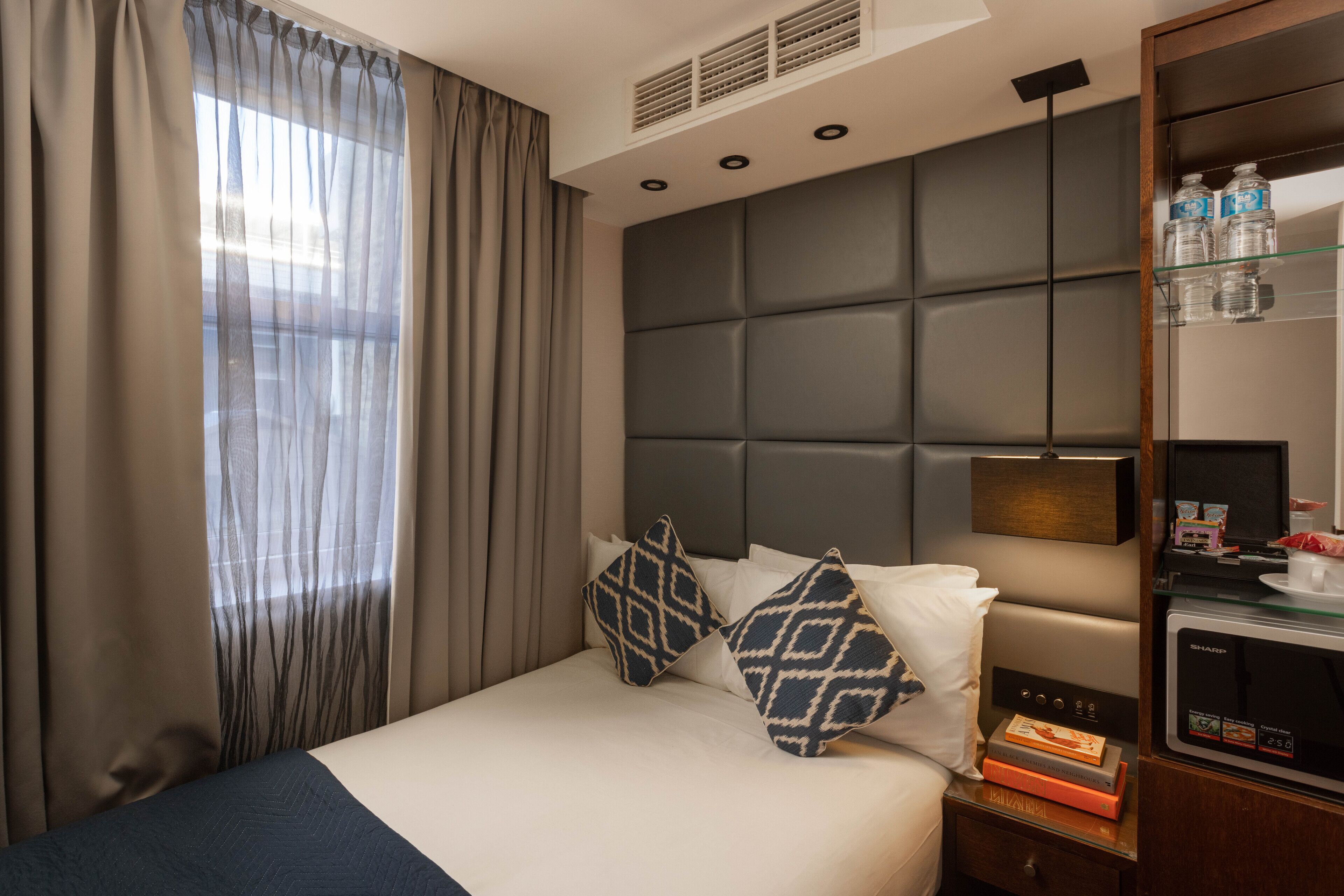 A hotel room with a bed, a nightstand, a bookshelf, and a window with curtains.