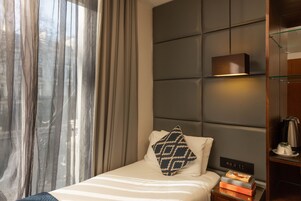 Hotellihuone, jossa on sänky, yöpöytä kirjoineen, toinen yöpöytä lampun kanssa ja suuri ikkuna, jossa on verhot.