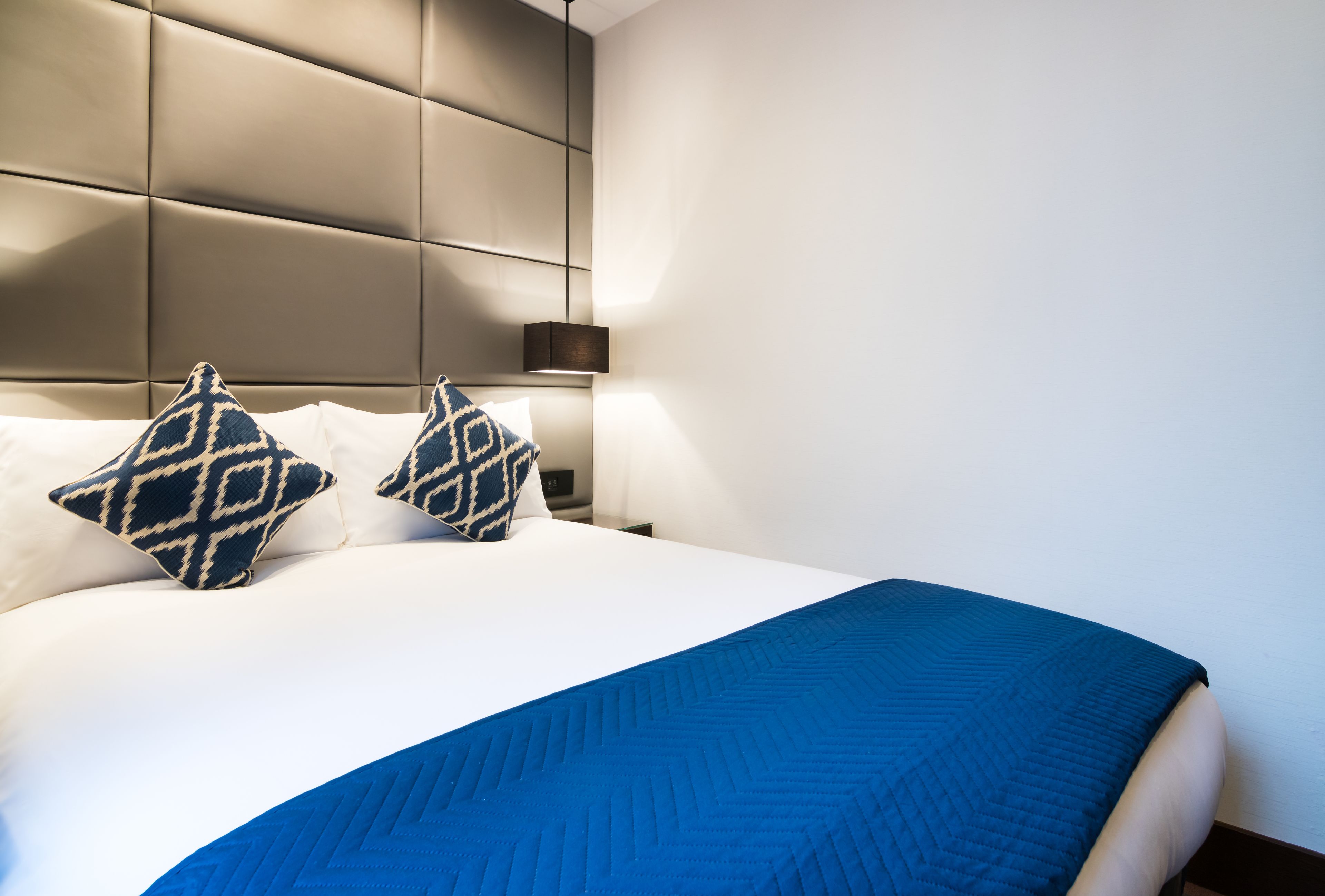 Een net opgemaakt bed met een blauwe deken en twee bedekte kussens, een modern hoofdbord en een nachtkastje met een lamp.