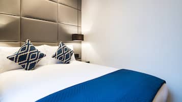 Een net opgemaakt bed met een blauwe deken en twee bedekte kussens, een modern hoofdbord en een nachtkastje met een lamp.