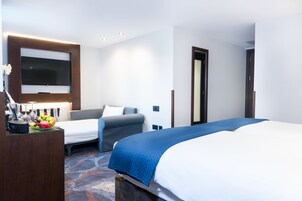 Hotellihuone, jossa on suuri sänky, pienempi sänky, työpöytä, jossa on litteänäyttöinen televisio, ja hedelmäkulho.
