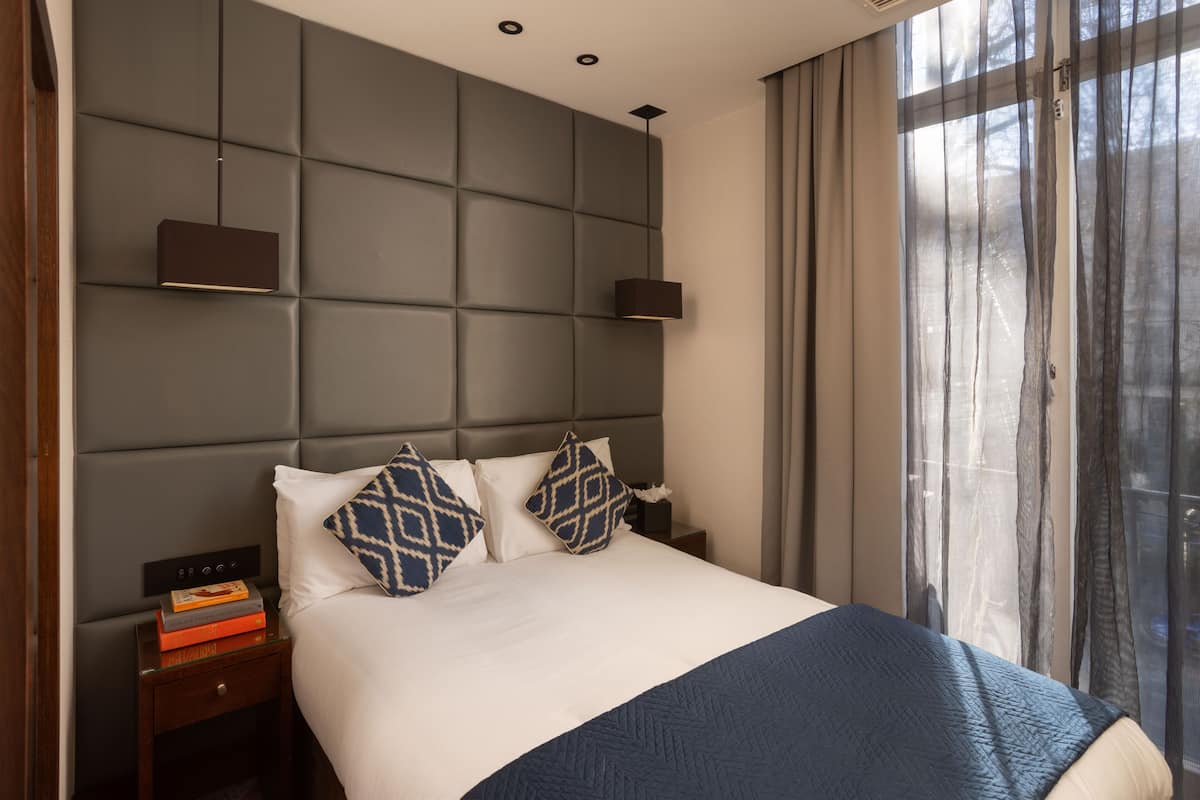 Una habitación de hotel moderna con una cama grande, un cabecero con respaldo acolchado, una mesita de noche con un libro y una ventana con cortinas translúcidas.