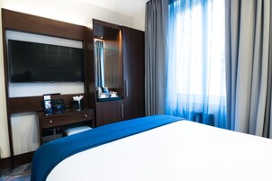 Ein Hotelzimmer mit einem großen Bett, einem Schreibtisch mit Flachbildfernseher, einem Spiegel und einem Fenster mit Vorhängen.