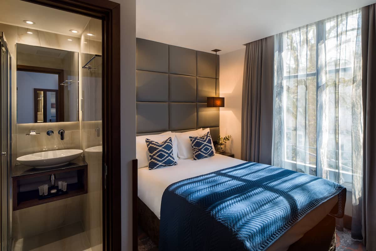 Una habitación de hotel moderna con una cama grande, una mesita de noche, un tocador, un espejo y un baño con lavabo y ducha.