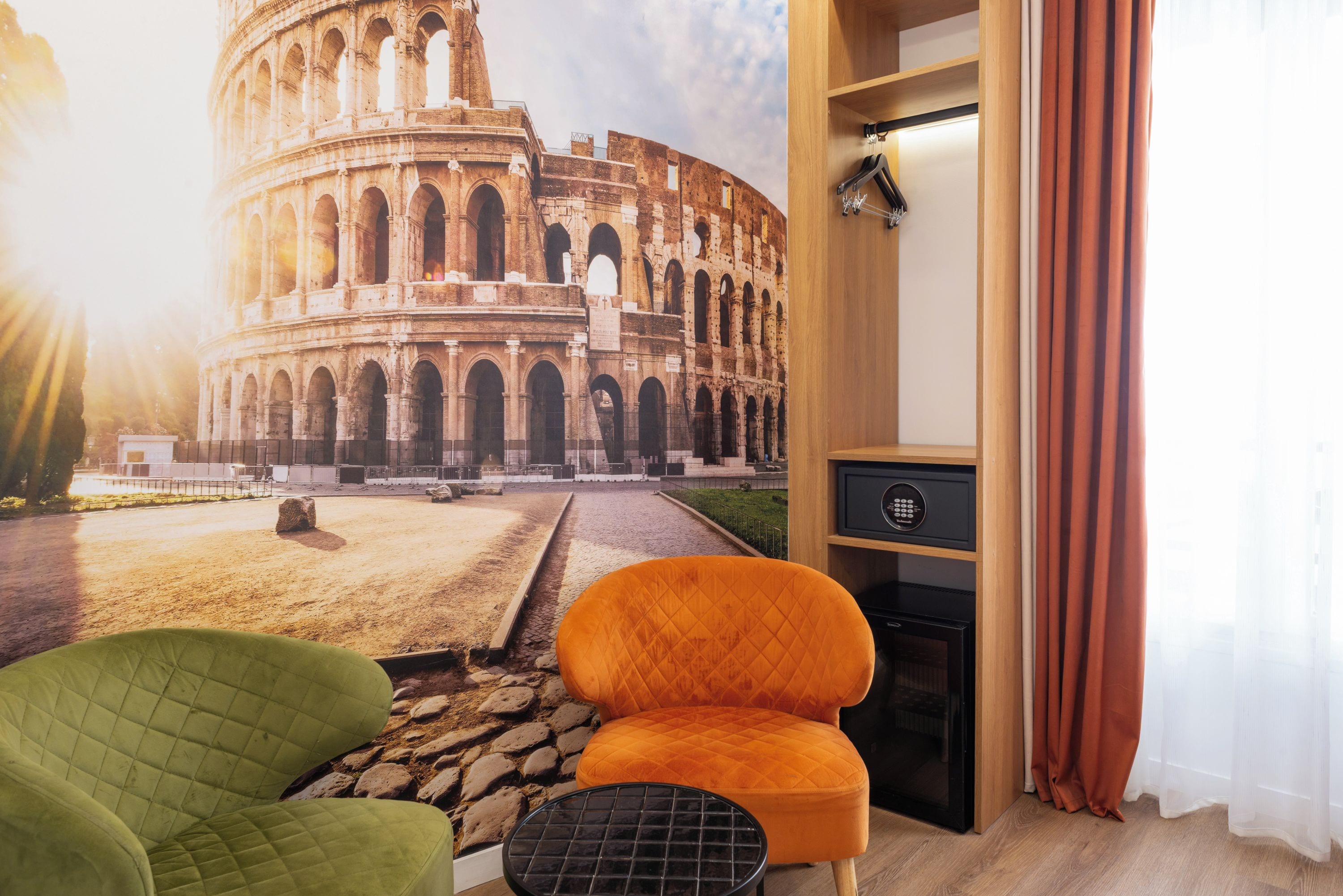 A room with a large window featuring a view of the Colosseum, a green armchair, an orange armchair, and a black coffee table.