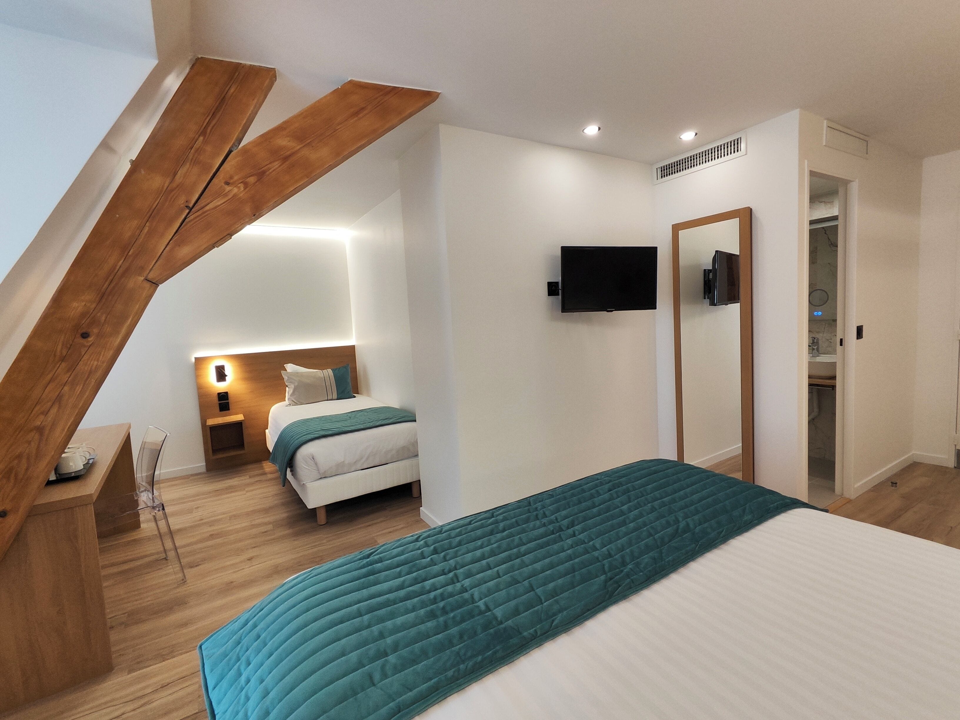 A modern bedroom with a sloped ceiling, a bed with a teal quilt, a bedside table, a TV mounted on the wall, and a walk-in closet.