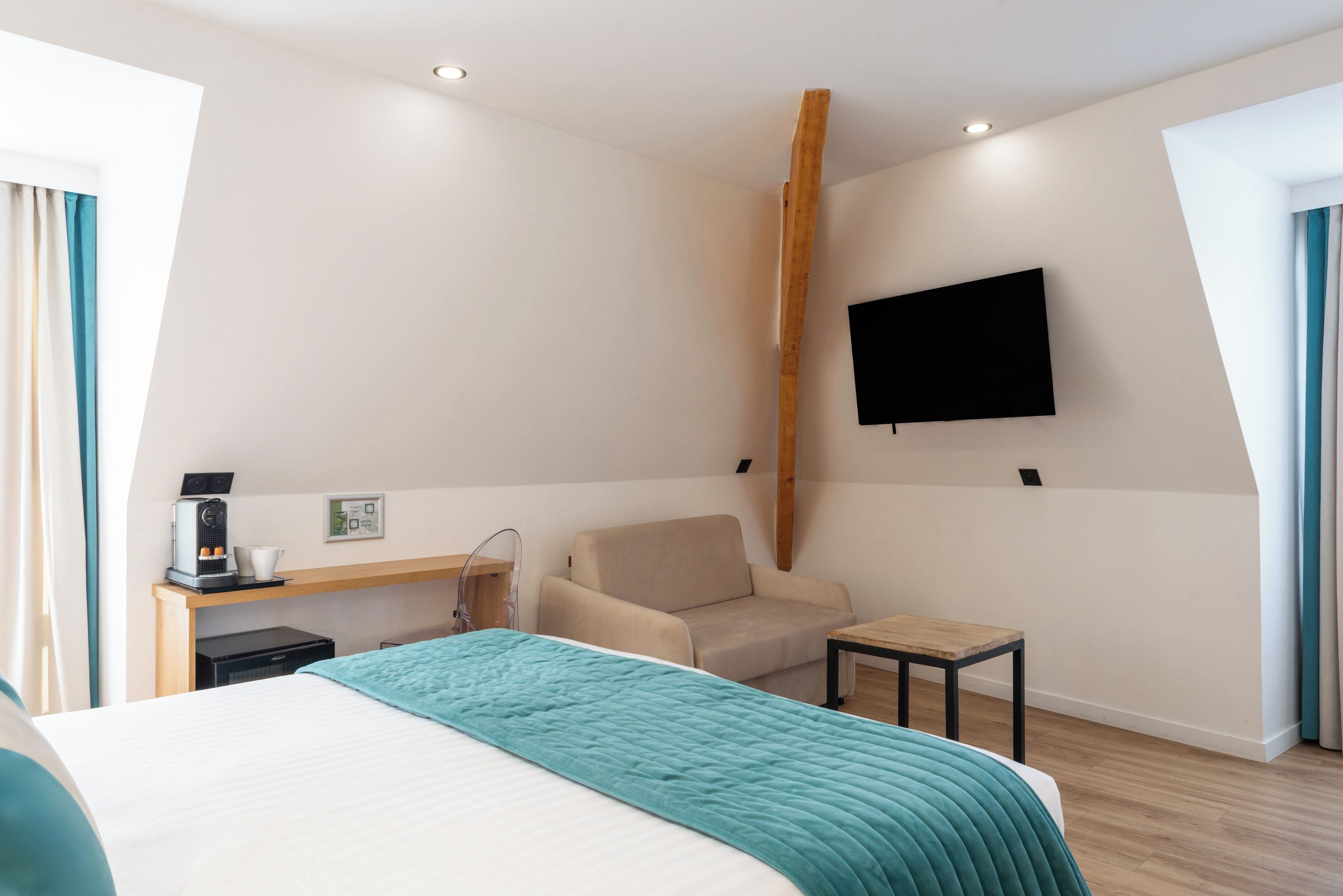 A hotel room with a bed, a desk with a coffee machine, a chair, a small table, and a TV mounted on the wall.