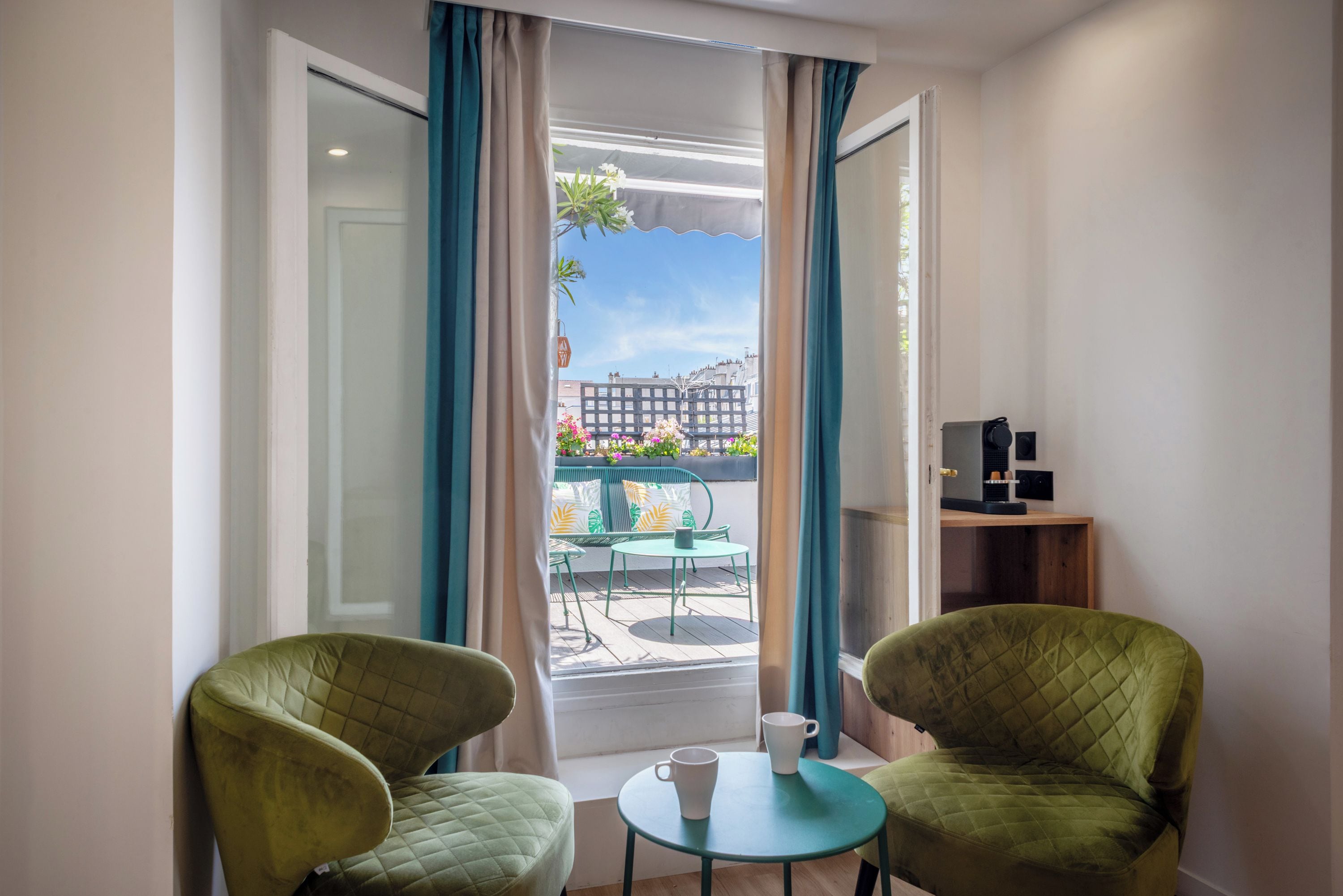 A room with two green armchairs, a small round table, and a view of a balcony with plants and a cityscape.