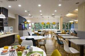 Un espace repas moderne avec des tables et des chaises rondes, un coin cuisine équipé d’un réfrigérateur et d’un évier, et de grandes fenêtres offrant une vue sur la verdure environnante.