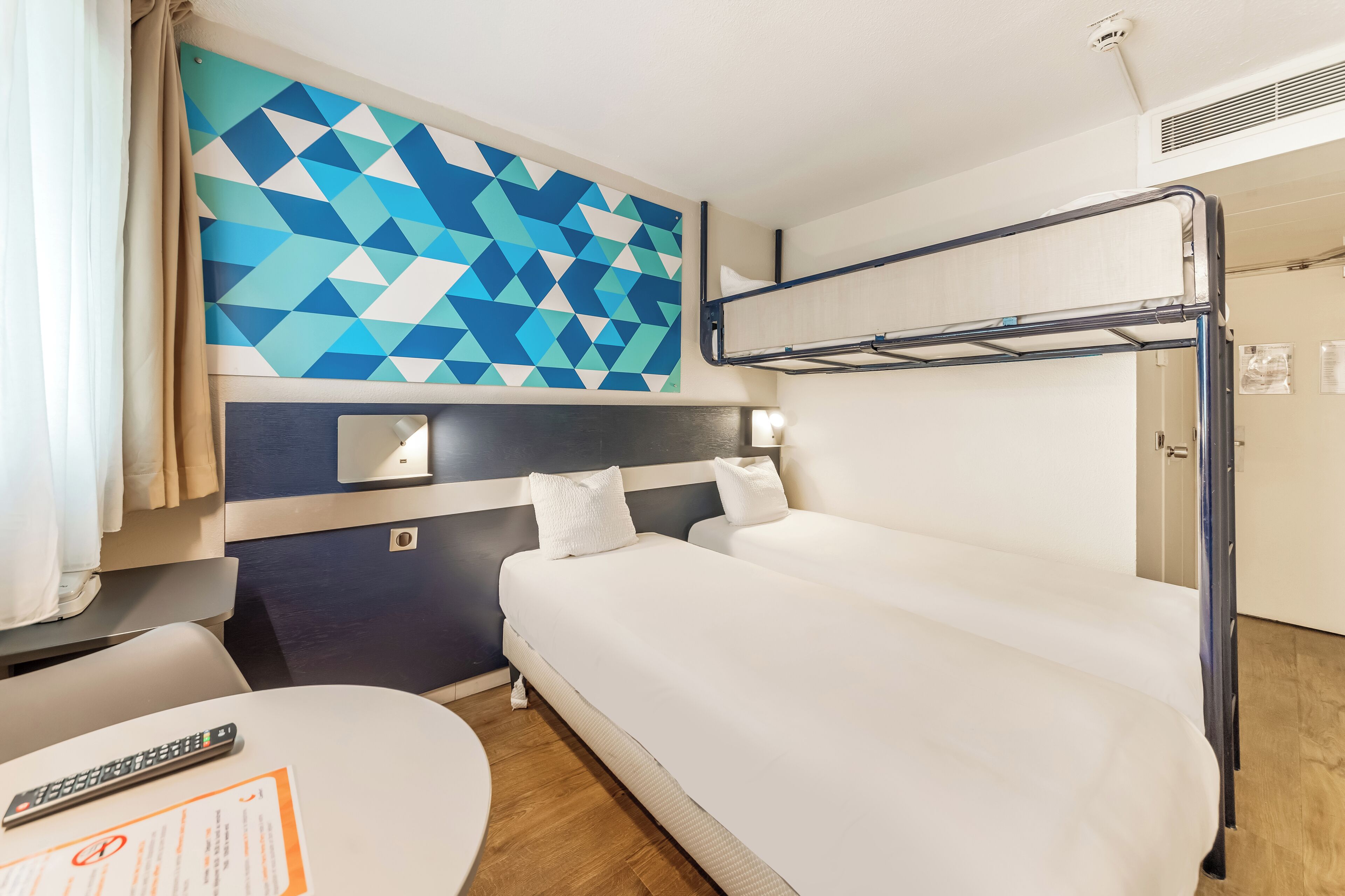 A hotel room with a bunk bed, a desk with a remote control, and a geometric-patterned wall.