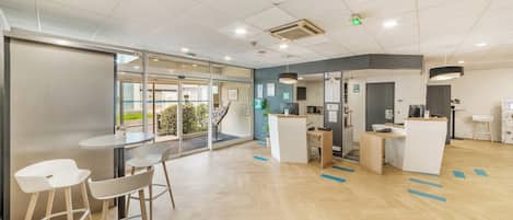 Uma área de recepção moderna com entrada por porta de vidro, balcão de recepção e um espaço de estar com mesas e cadeiras.