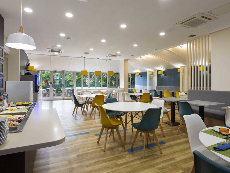 A modern dining area with round tables and chairs, a buffet with food items, and a view of the outdoors through large windows.