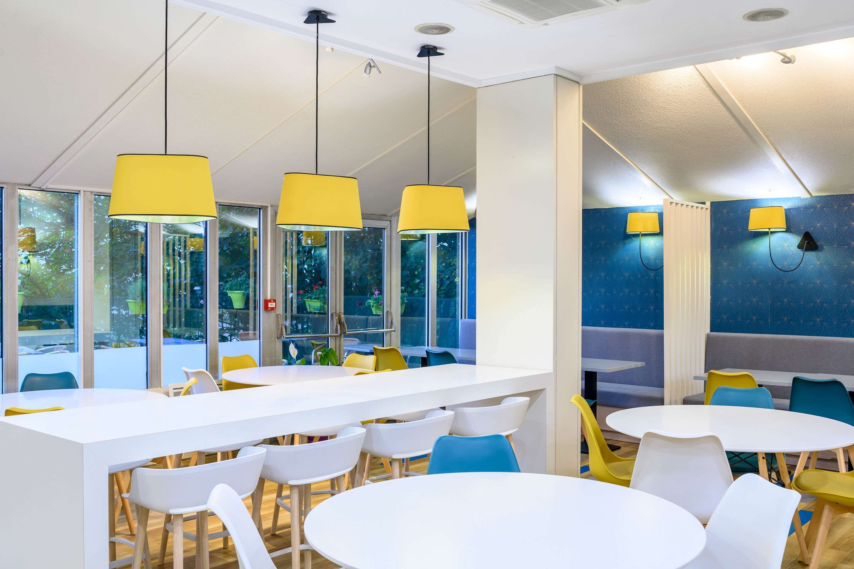 A modern indoor space with white tables and chairs, yellow pendant lights, and large windows with a view of greenery.