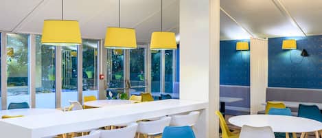 A modern indoor space with white tables and chairs, yellow pendant lights, and large windows with a view of greenery.