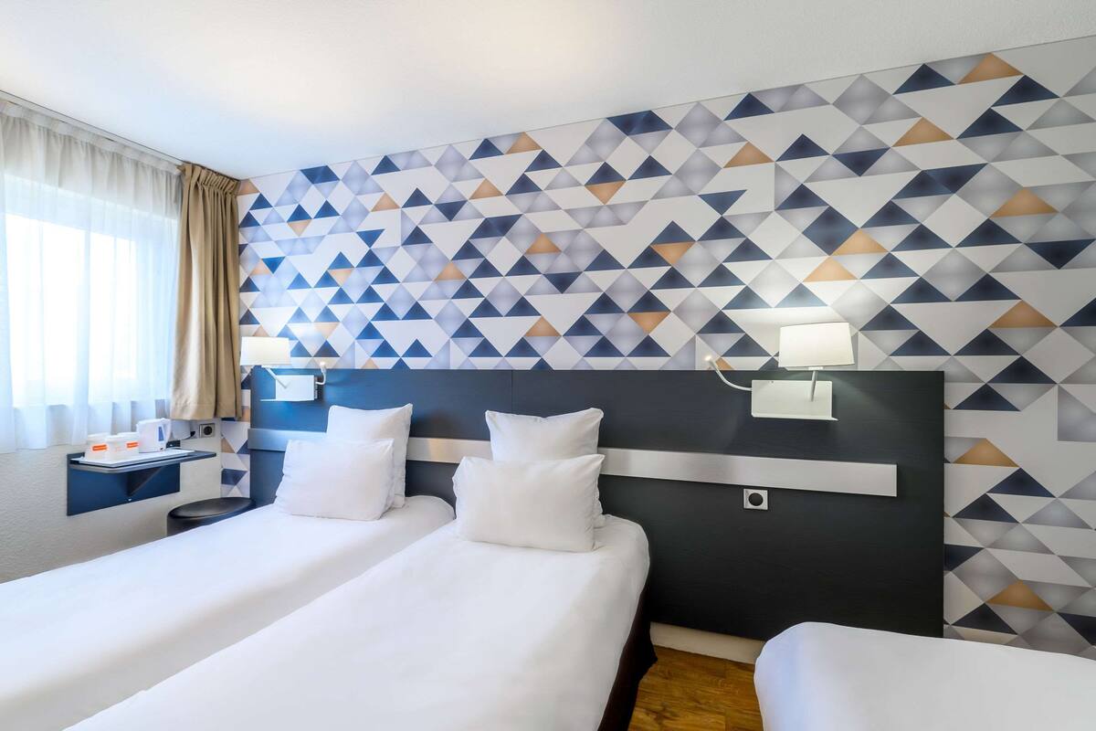 A hotel room with two beds, a nightstand, a wall-mounted lamp, and a geometric-patterned wall.