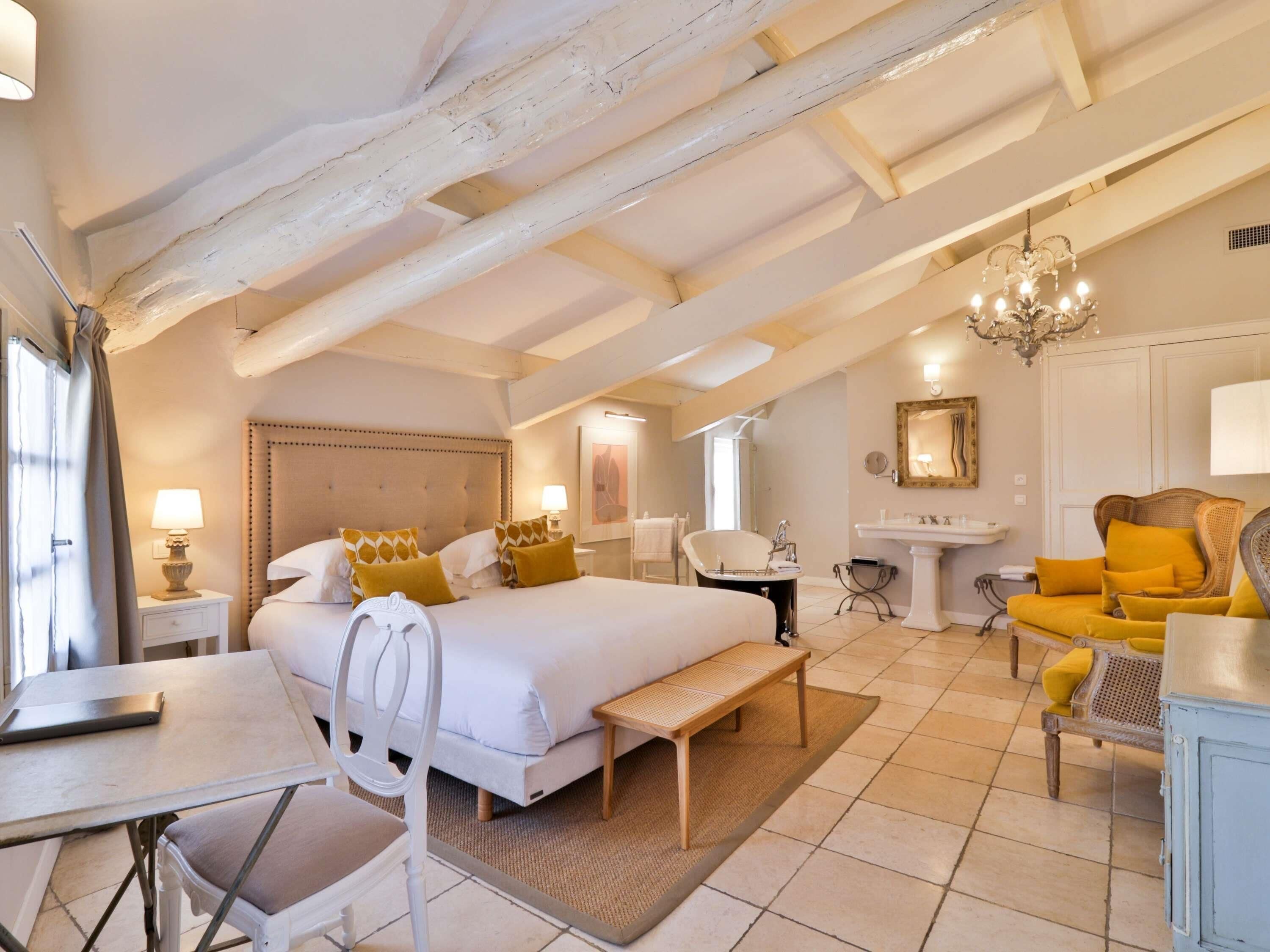 A bedroom with a bed, bedside table, lamp, chair, bench, and a view of the bathroom.