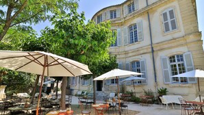 An outdoor café area with tables and chairs, surrounded by trees and a historic building with multiple windows and shutters. - Chateau de Mazan - Handwritten Collection (Mazan)