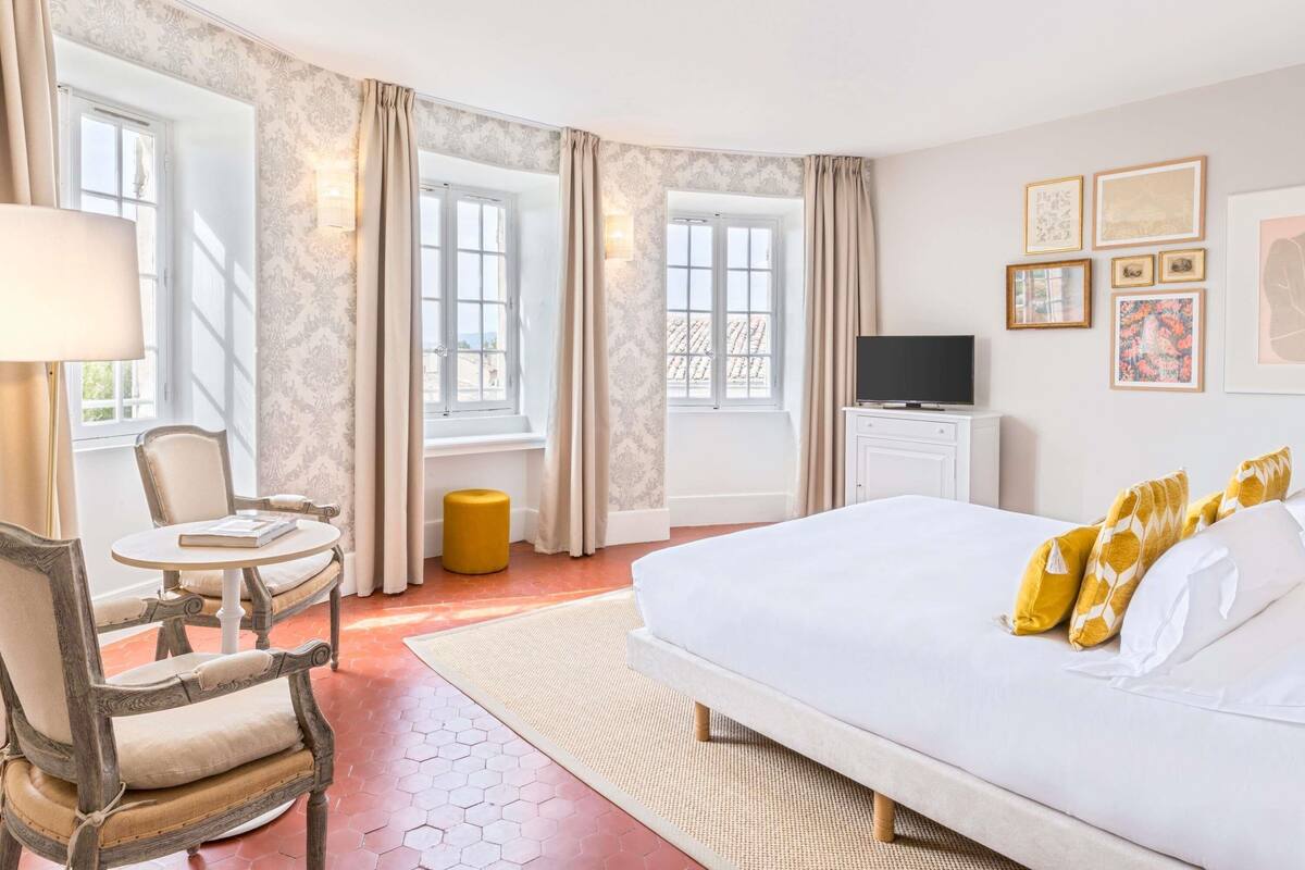 A hotel room with a bed, a chair, a small table, a TV, and artwork on the wall.