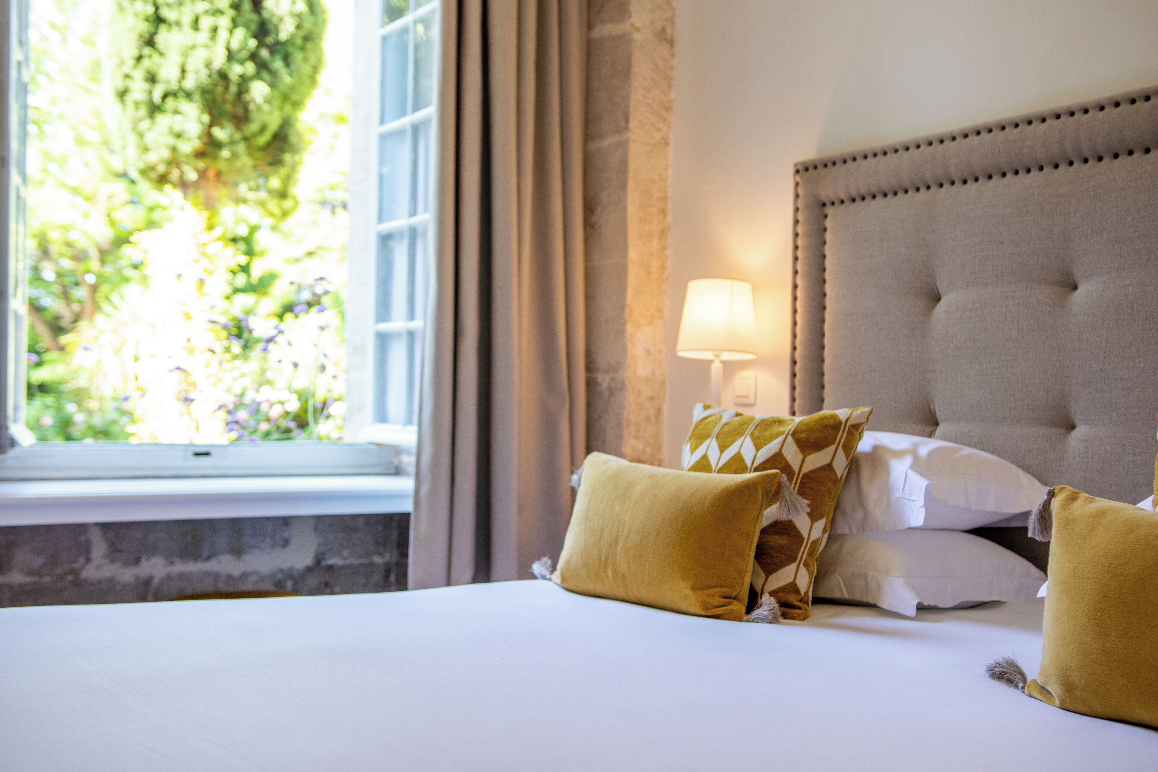 Una cama bien hecha con sábanas blancas y dos almohadas amarillas, una lámpara de noche y una ventana con cortinas.