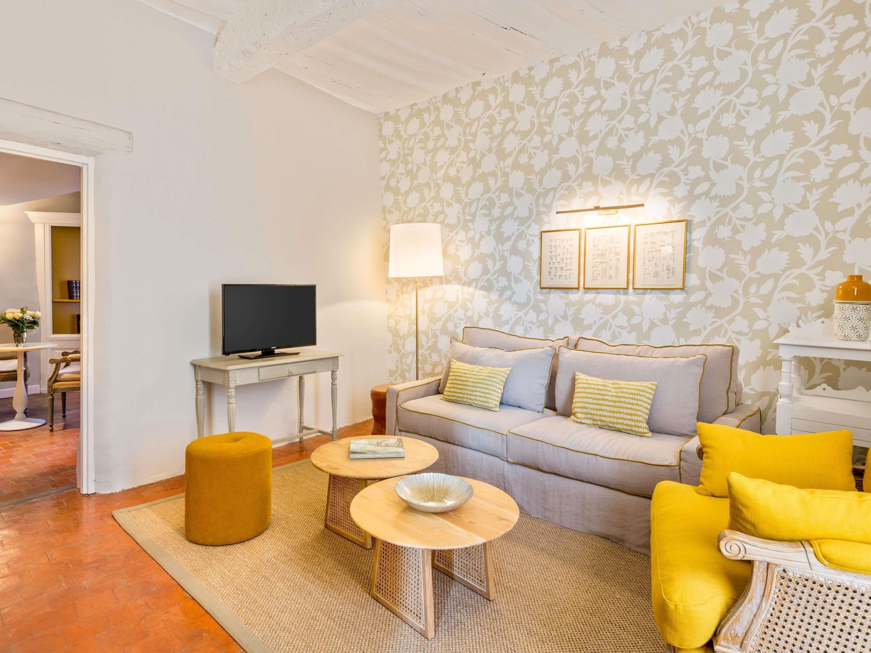 Ein Wohnzimmer mit einer Couch, einem gelben Sessel, zwei kleinen Tischen, einem Fernseher und einer Wand mit Muster.