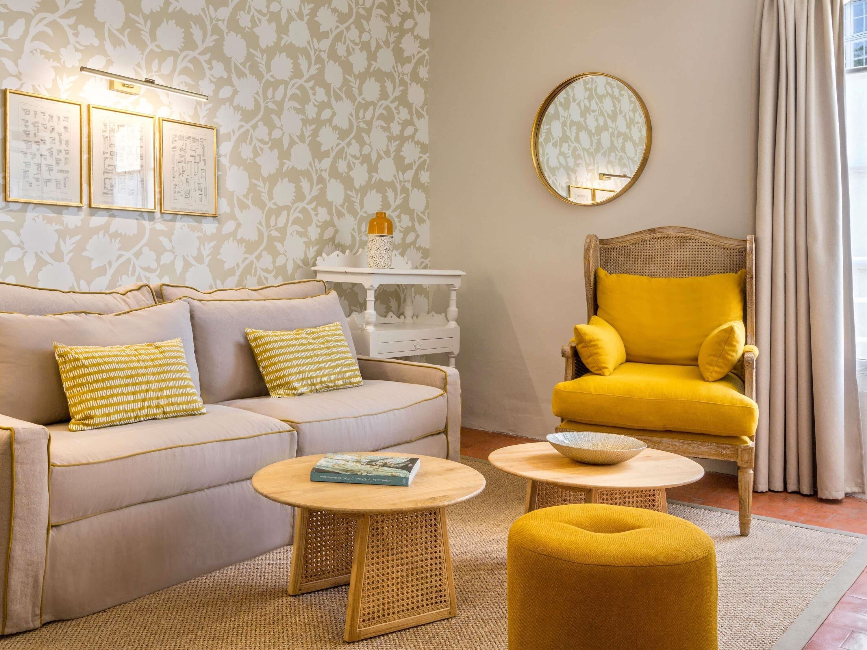 A living room with a sofa, armchair, round coffee table, and ottoman. There is a wall-mounted mirror and framed artwork.