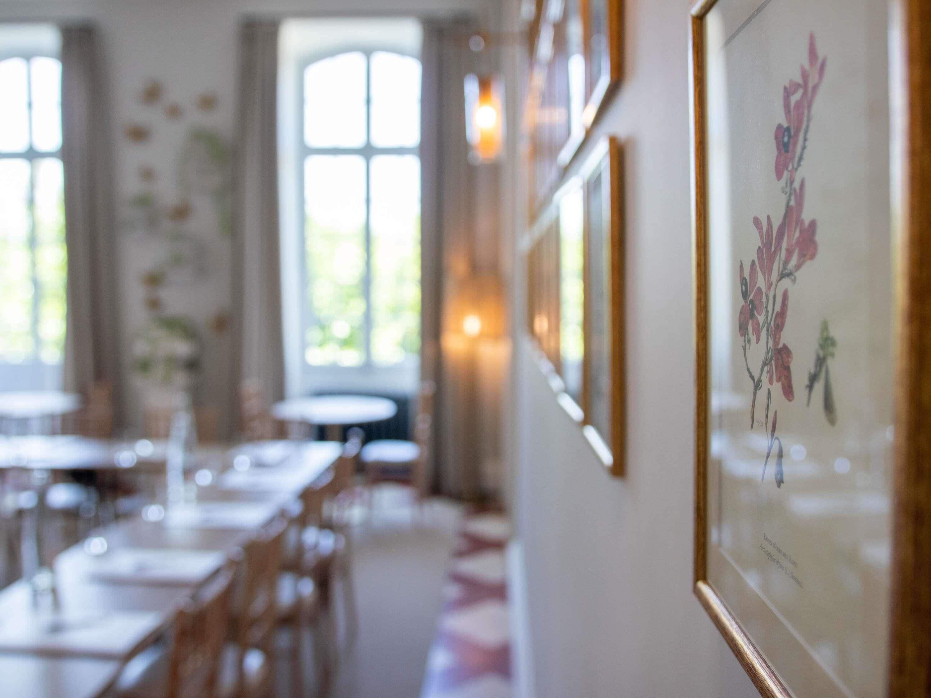 A dining area with tables and chairs, framed artwork on the wall, and large windows allowing natural light.