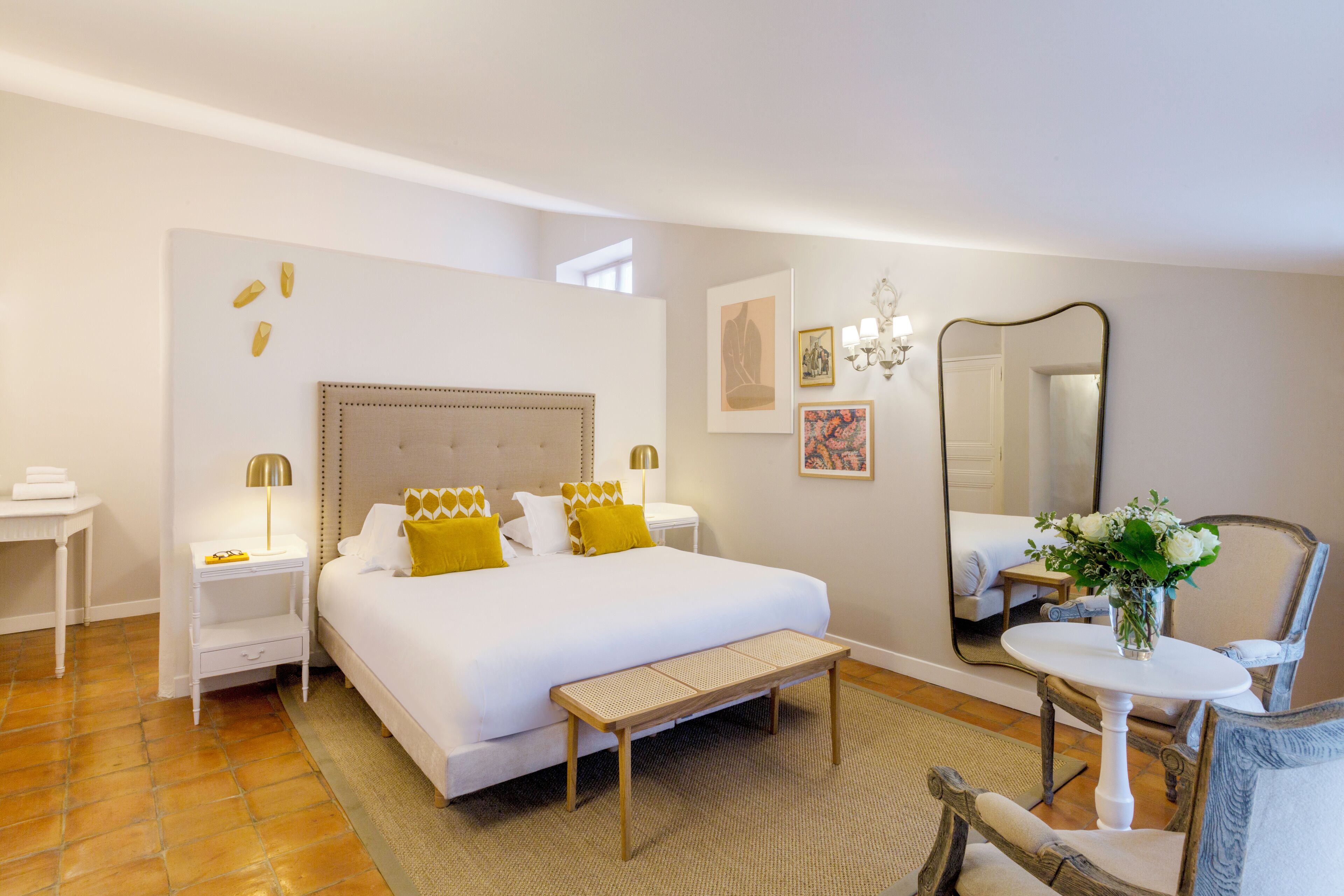 Ein Hotelzimmer mit einem großen Bett, Nachttischen, einer Bank, einem kleinen Tisch mit einer Vase, einem Sessel und einem Spiegel.