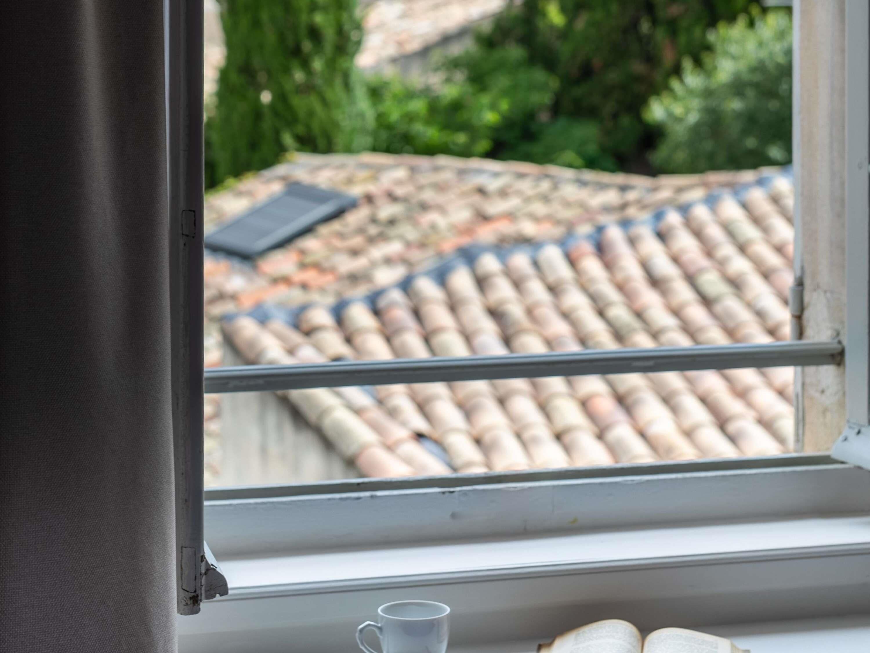Une fenêtre donnant sur un toit recouvert de tuiles et une petite table avec une tasse.