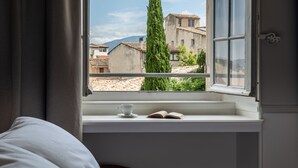 A bedroom with a bed, a window with a view of buildings and a tree, a cup, and an open book on the windowsill. - Chateau de Mazan - Handwritten Collection (Mazan)