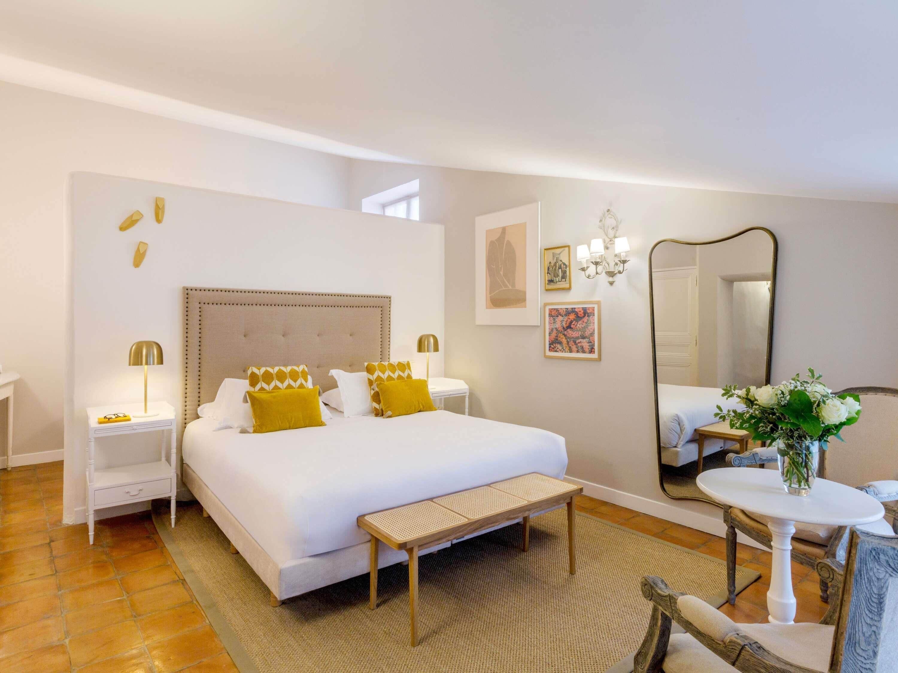 A hotel room with a large bed, bedside tables, a small round table, a bench, a mirror, and artwork on the walls.