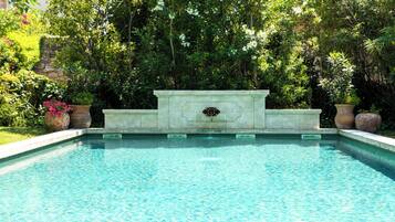 Piscina rectangular con un borde de piedra y una fuente decorativa en el centro, rodeada de abundante follaje y plantas en macetas.