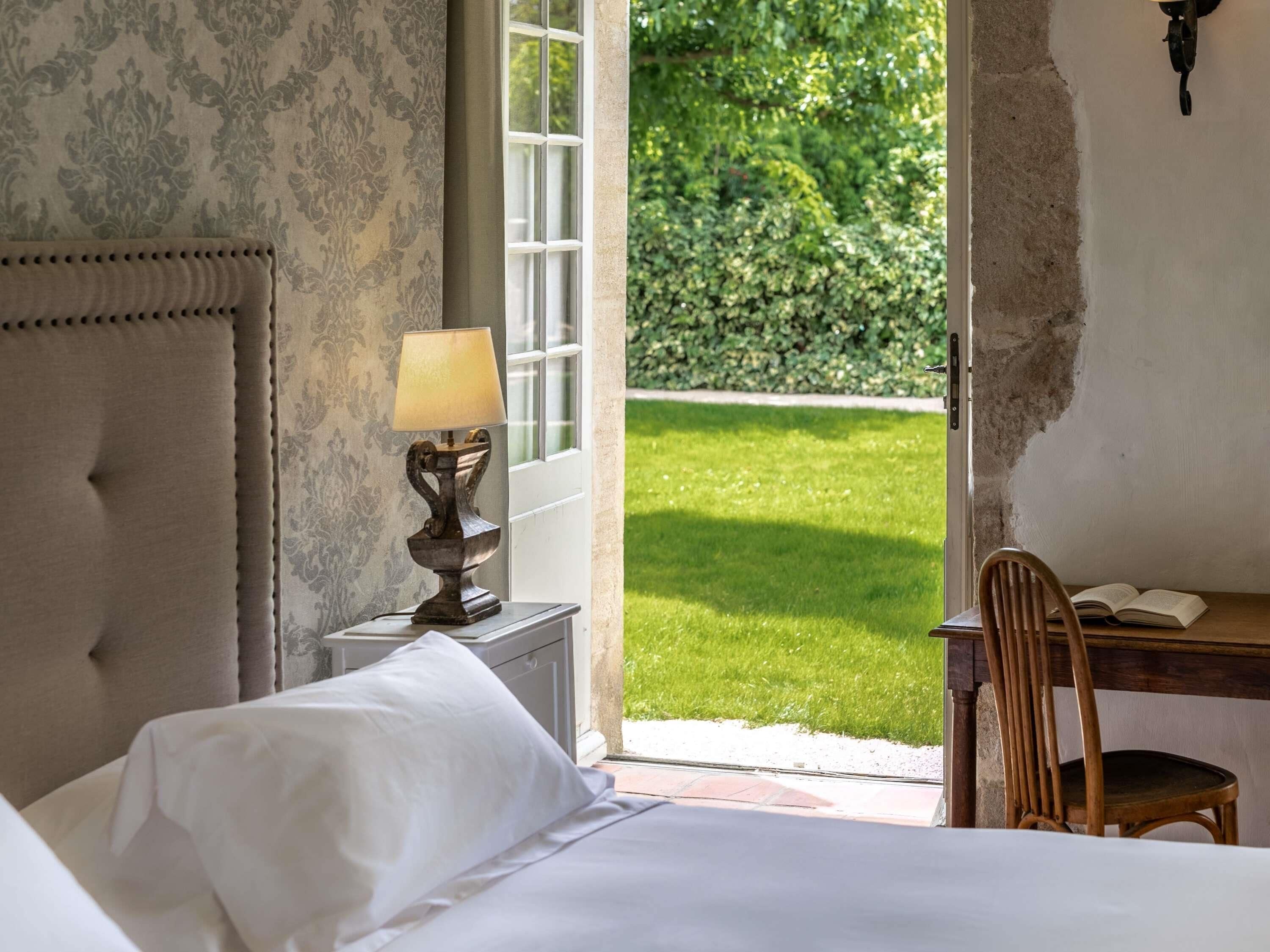 A bedroom with a bed, a bedside table with a lamp, a chair, and a view of a garden.