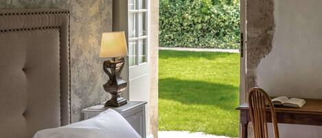 A bedroom with a bed, a bedside table with a lamp, a chair, and a view of a garden.