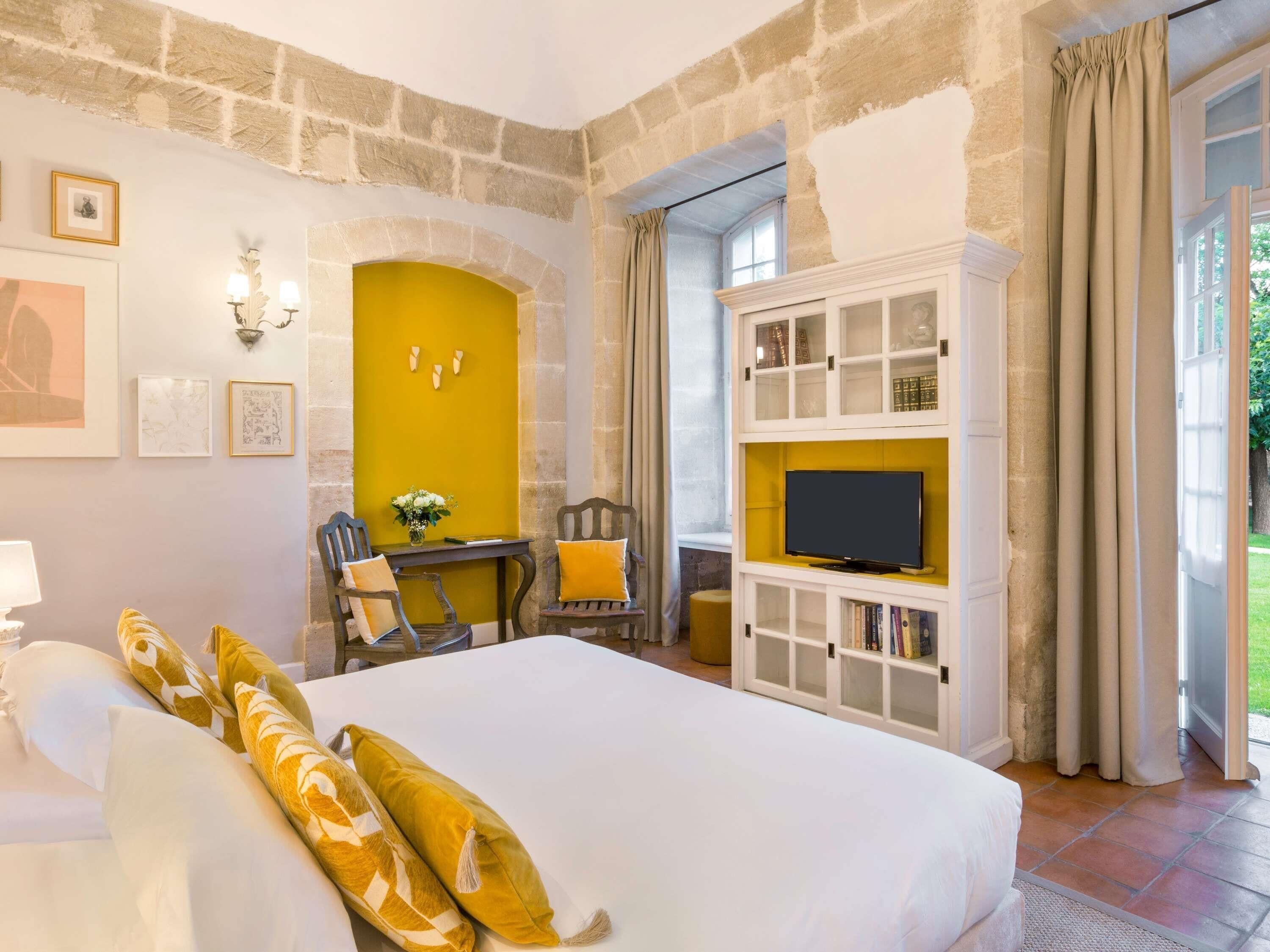A hotel room with a large bed, a television on a white cabinet, a desk with a chair, and a stone wall.