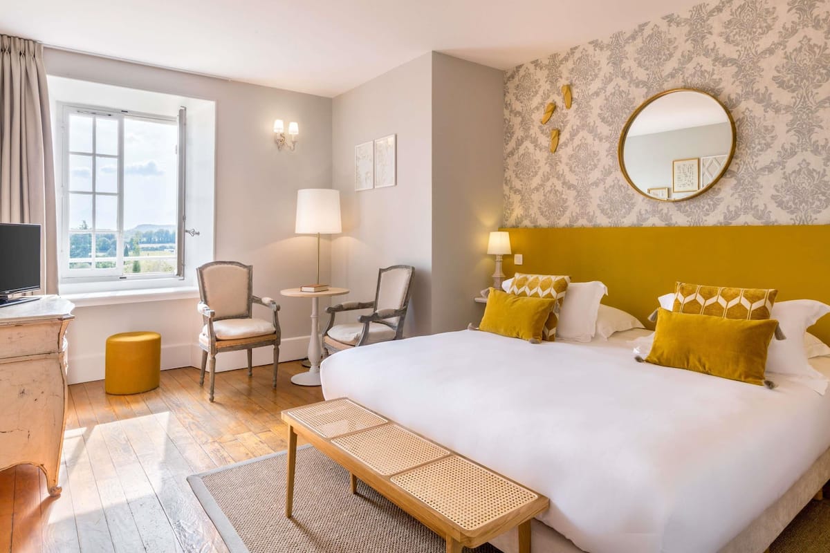 A hotel room with a bed, a desk, a chair, a small table, a window, and a wall with a mirror and framed pictures.