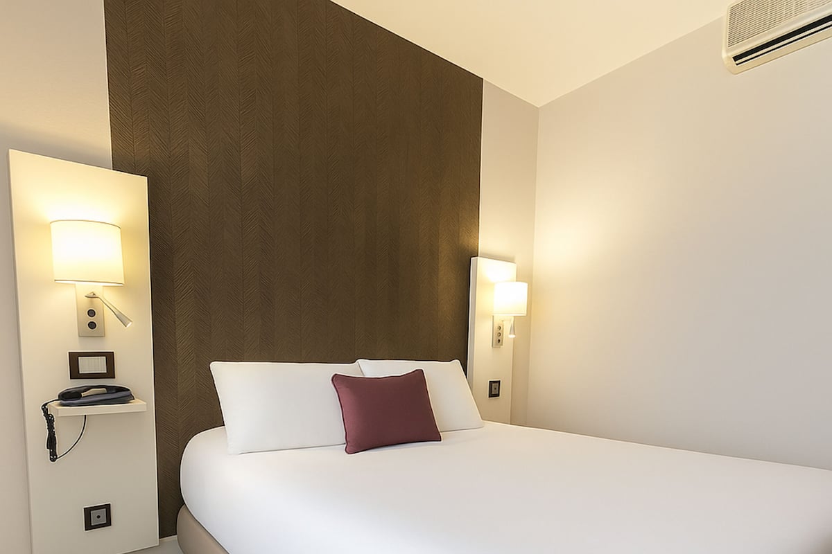 Habitación de hotel con una cama grande, dos lámparas de noche, un calentador de pared y una mesita con una lámpara.