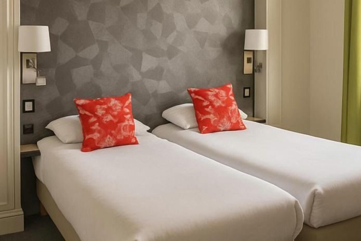 A hotel room with two beds, each with a white bedspread and a red pillow. There are two bedside lamps and a wall-mounted light.