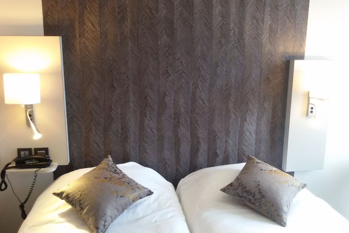 Una habitación de hotel con dos camas, una mesita de noche con lámpara y un papel tapiz estampado.